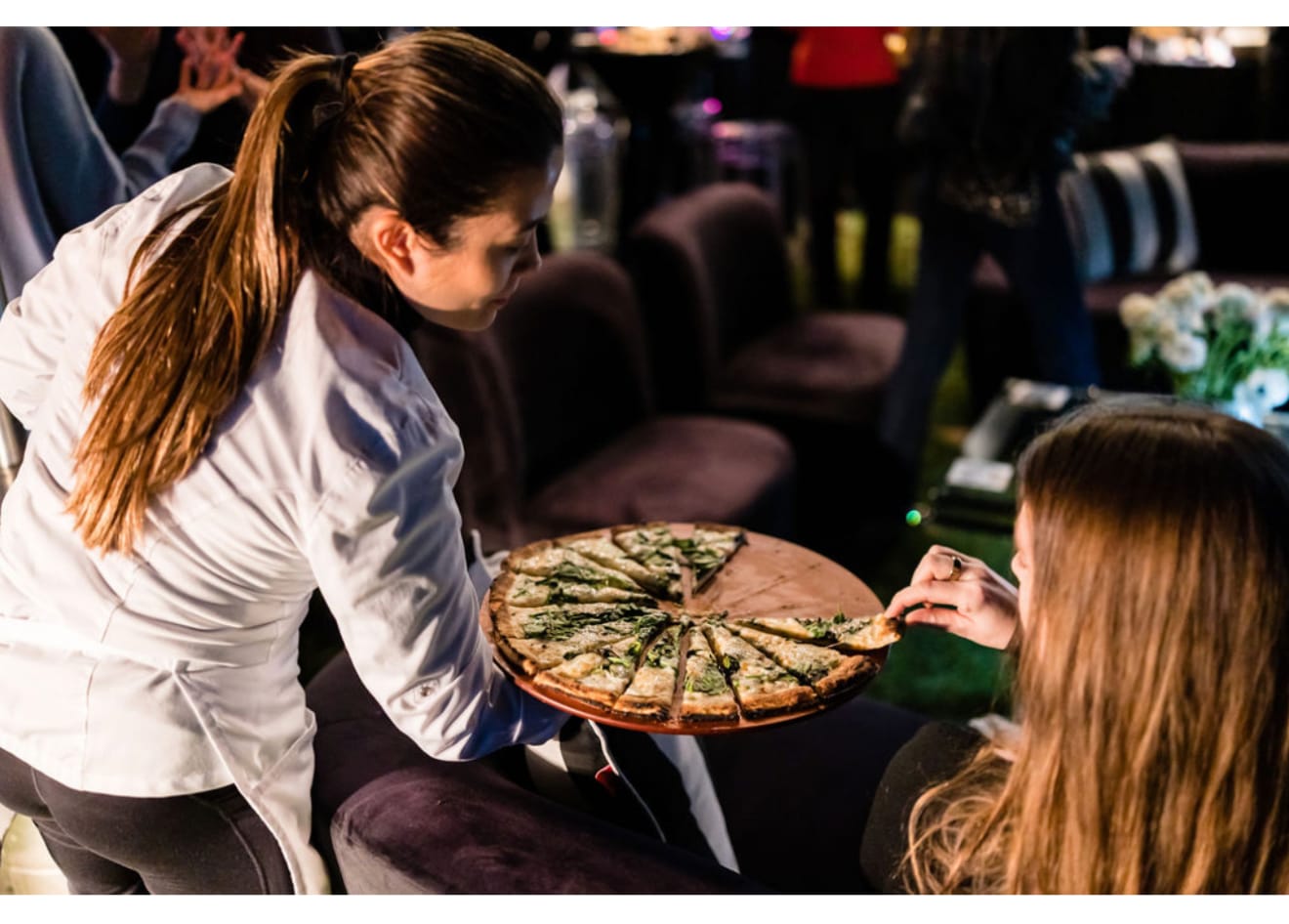 Server presenting a freshly sliced pizza to a seated guest at an evening Orange County event